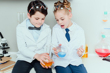 Schoolchildren in white coats holding reagents in flasks sitting in science laboratory