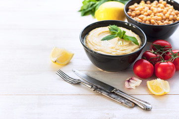 Bowl with hummus, chickpeas, lemon, cherry tomatoes and herbs on a white rustic wooden background. Middle Eastern cuisine. Background hummus with place for text.