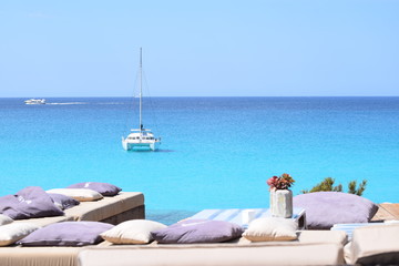 Luxuriöse blaue Yacht ankert im türkisfarbenen Wasser vor der Küste von Formentera, im Vordergrund Bänke und Kissen am Ufer, im Hintergrund ein Motorboot - obrazy, fototapety, plakaty