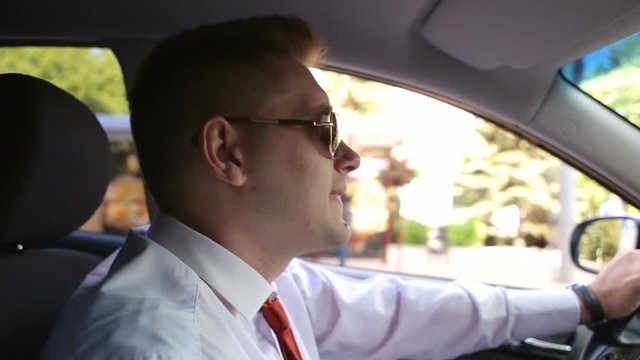 Happy businessman in a shirt and sunglasses is driving a modern car and singing. Cheerful businessman sings behind wheel of a car.
