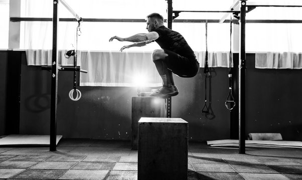 Fit Young Man Jumping Onto A Box As Part Of Exercise Routine. Man Doing Box Jump In The Gym. Athlete Is Performing Box Jumps