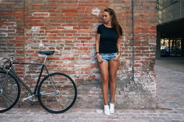An outdoor portrait of a young cute student girl wearing black blank t-shirt and blue jeans shorts with a fixed gear bicycle while standing on the brick wall background. Empty space for text or design