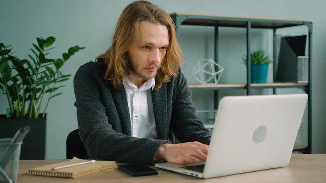 Young man sitting in the office and typing on his laptop computer. Long hair hipster using laptop pc, looking at screen while sitting at the office table.