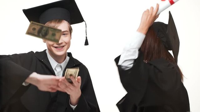 Happy young Caucasian male throwing dollars at camera while African American female in black robe and square academical cap rejoicing smiling near him. Footage on white background in slowmotion
