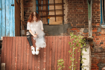 Beautiful vape teenager. Portrait of a pretty young white girl with red curly hair vaping an...