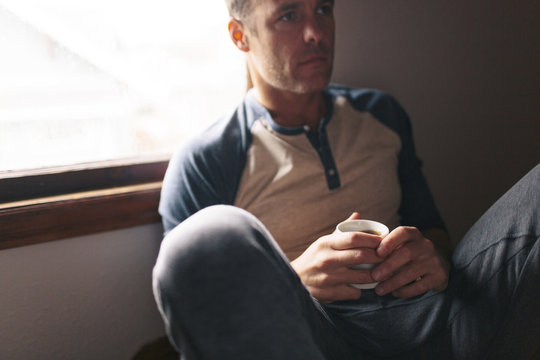 Thoughtful man with coffee cup sitting by window at home