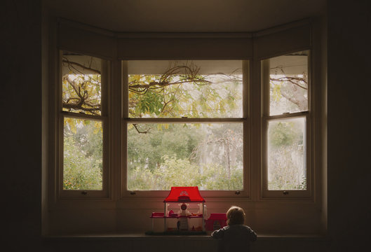 Rear view of baby girl playing with dollhouse at bay window in house