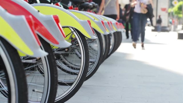 close up perspective view of bicycle rear wheels and lights parked in city bike stand with people walking on the sidewalk in the background