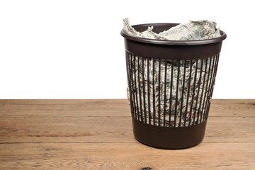 Garbage basket full of crumpled dollars stands on an old wooden table isolated on a white background