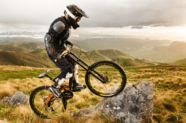 Plakat Ryder in full protective equipment on the mtb bike climbs on a rock against the backdrop of a mountain range and low clouds