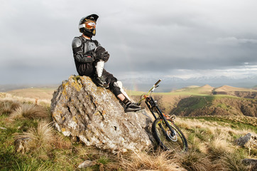 A professional downhill rider sits on a stone resting after a tense race on a mtb bike