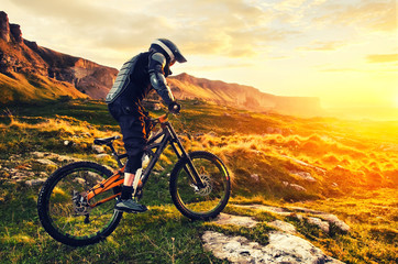 The rider in full protective equipment on the mtb bike is riding toward the sunset in the rays of...