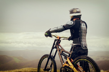 Fototapeta na wymiar The rider in the full-face helmet and full protective equipment on the mtb bike stands on a rock against the background of a ridge and low clouds