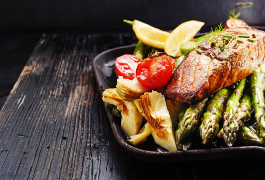 Recipe - delicious portion of grilled salmon fillet with asparagus, tomatoes, artichoke, spices on a black wooden board. Close up, copyspace, selective focus.