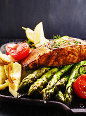 Recipe - delicious portion of grilled salmon fillet with asparagus, tomatoes, artichoke, spices on a black wooden board. Close up, copyspace, selective focus.