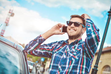 Inviting friend for a journey. Handsome young man talking on the mobile phone and smiling while leaning at his car