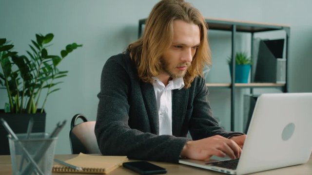 Young man sitting in the office, talking on the phone and typing on laptop computer. Long hair man looking at screen, checking information while talking on the cell phone.