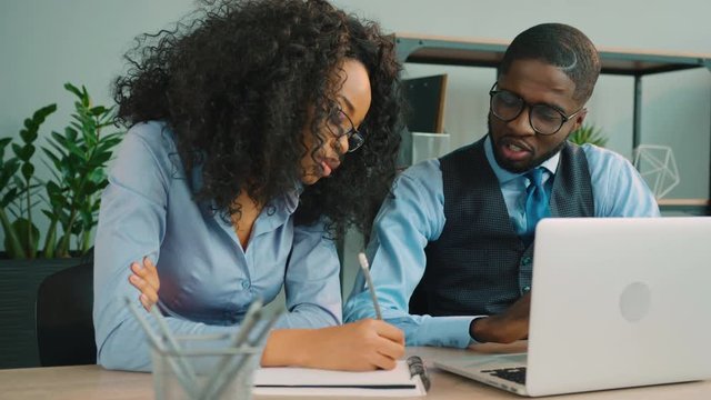 Team of two african american business people working together in in the office. African American businessman and businesswoman talking, using laptop and writing down information.