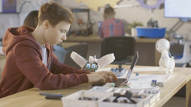 Smart Young Boy Works on a Laptop For His New Robot Programing Project in Computer Science Class. Other Children Learning in the Background. Shot on RED EPIC-W 8K Helium Cinema Camera.