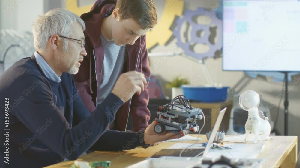 Poster in computer science class teacher examines programed robot engineered by his student for school proj
