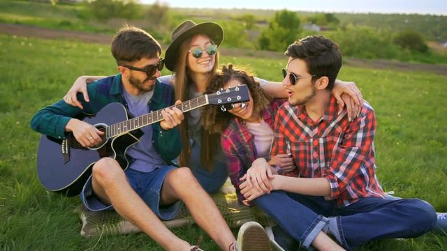 Four friends are embracing, plays guitar and sings songs outdoors at sunset. They very happy and cheerful