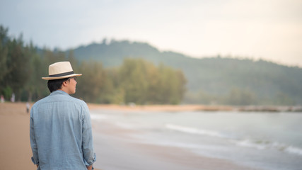 Young Asian man with jean shirt and hat walking on beautiful tropical beach and see the sunset, happy vacation time and summer holiday travel concepts