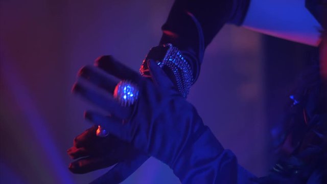 Woman dressed in the style of a Great Gatsby with long silk gloves shows her rings and bracelet on party. Neon blue and red light.