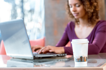 Fototapeta premium Urban vibes. Selective focus on a paper cup on the table young African female student studying on her laptop