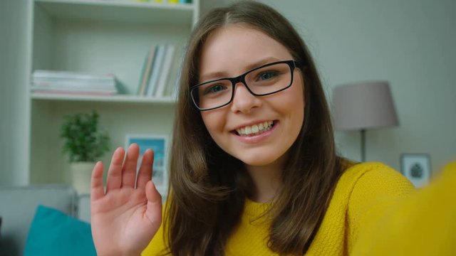 Beautiful young teen girl video chatting, waving hand, blogging in the living room at home. Girl smiling to the camera. Close up