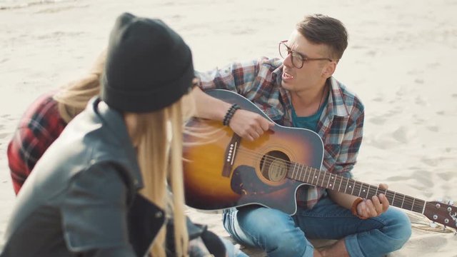 Two blondes and a guy in a plaid shirt sing songs under a guitar while sitting on a log on the sea coast during sunset close-up.