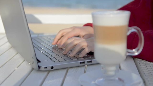Young woman typing on her laptop in cafe on the beach with cup of coffee