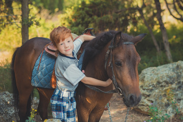 Cute red hair boy smiling to camera and standing in forest with horse