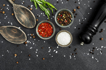 On a black stone surface of spices, black pepper mill, wooden spoons, in glass bowls pepper peas, salt, paprika, rosemary