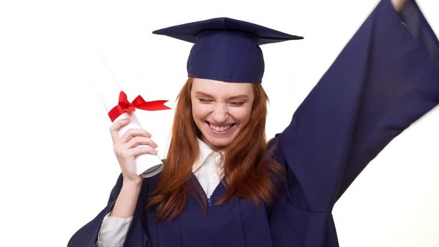 Exciting Caucasian ginger graduate girl in blue robe and square academical cap showing diploma scroll and rejoicing in white background