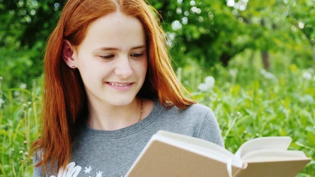 Red-haired teenage girl reading a book in the park. 4K slow motion video