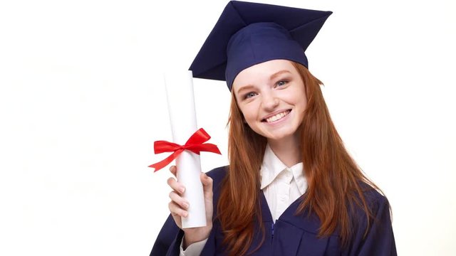 Smiling ginger young graduate girl in blue robe and square academical cap entering frame holding her diploma. Footage on white background