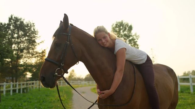 SLOW MOTION, CLOSE UP: Two cheerful young girls sitting on strong chestnut gelding and embracing it. Pretty girlfriends bareback riding stunning brown stallion on horse ranch at gorgeous summer sunset