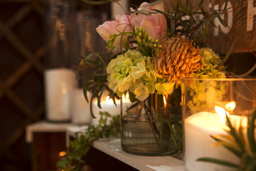 Romantic decor: glass vase with flowers in it, lit candles around it. Dim lights from candles only. Night time. Rich interior. Warm toning