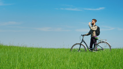 A young cyclist drinks water. Standing beside his bicycle on a green hill against a blue sky.
