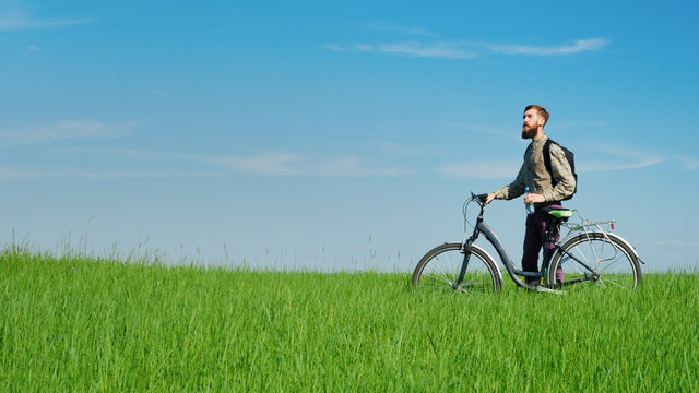 A young cyclist drinks water. Standing beside his bicycle on a green hill against a blue sky. An idyllic landscape - Powered by Adobe
