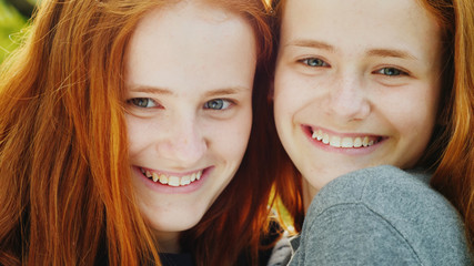 Close-up Portrait of two happy sisters twins. They are red-haired, look at the camera, smile, hug. The sun beautifully illuminates their hair