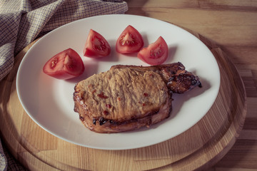 Pork steak and sliced tomato on a white plate on a wooden round tray, vintage effect
