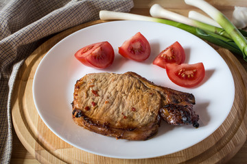 Pork steak, sliced tomato on a white plate, green young shoots of garlic on a wooden tray, shallow depth of field