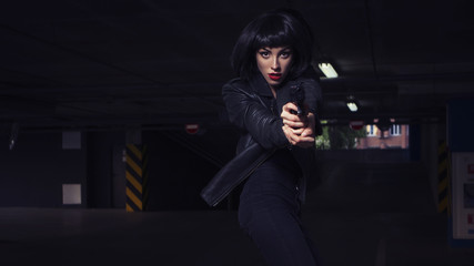 Beautiful brunette sexy spy agent (killer or police) woman in leather jacket and jeans with a gun in her hand running after someone, to catch him on parking