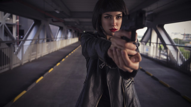 Beautiful brunette sexy spy agent (killer or police) woman in leather jacket and jeans with a gun in her hand running after someone, to catch him on the bridge road