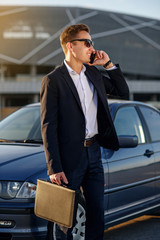 Attractive handsome businessman with diplomat talking on smartphone near the car