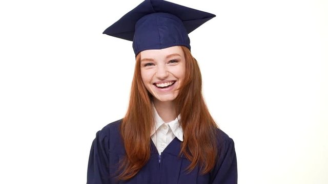 Happy beautiful young foxy graduate girl in blue robe and square academical cap laughing sweetly on white background in slowmotion