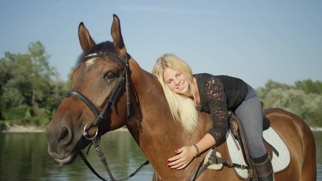SLOW MOTION, CLOSE UP, DOF Smiling young female rider caressing and hugging her dark brown mare on stunning sunny summer day at the riverside. Portrait of a beautiful Caucasian woman embracing a horse