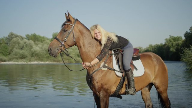 SLOW MOTION, CLOSE UP, DOF: Young smiling blonde girl mounted on beautiful brown stallion standing in shallow water in the river on sunny summer day. Caucasian woman embracing her beautiful horse