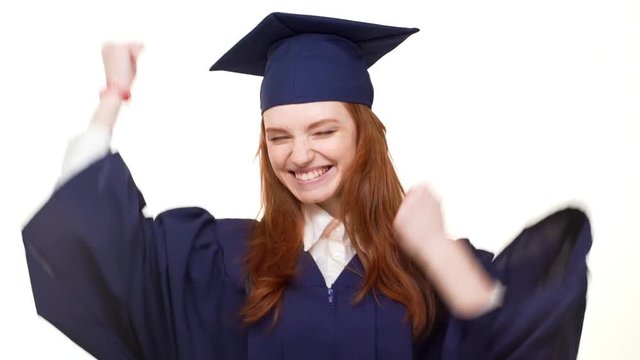 Young attractive Caucasian redhead graduate girl in blue academical square cap and robe rejoicing smiling laughing on white background in slowmotion